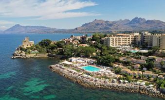 a resort with a large pool and a mountainous backdrop is nestled in a rocky coastline at Domina Zagarella Sicily