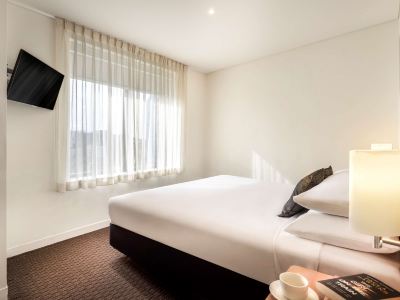 a neatly made bed is in a room with a window and a television on the wall at Ibis Melbourne Hotel and Apartments