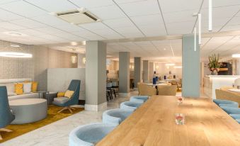 a modern hotel lobby with a wooden reception desk and several chairs arranged around it at NH Amsterdam Schiphol Airport