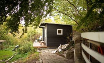 a small black house surrounded by trees , with a path leading up to it , and a wooden deck on one side at Paradiset