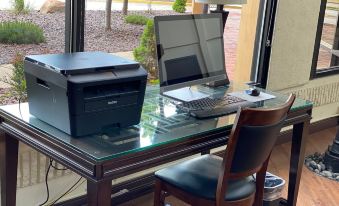 a glass desk with a laptop , printer , and keyboard is situated next to a window at The Atrium Hotel on Third