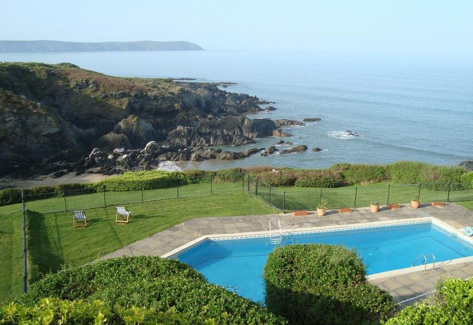 a large swimming pool is surrounded by a well - maintained lawn and overlooks the ocean with a rocky coastline in the background at Watersmeet Hotel