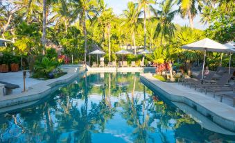 a large outdoor pool surrounded by palm trees , with lounge chairs and umbrellas placed around the pool at Castaway Island Fiji