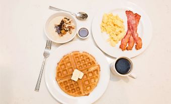 a breakfast table with a plate of waffles , bacon , scrambled eggs , and sausage , accompanied by a cup of coffee at Staybridge Suites Denver North - Thornton