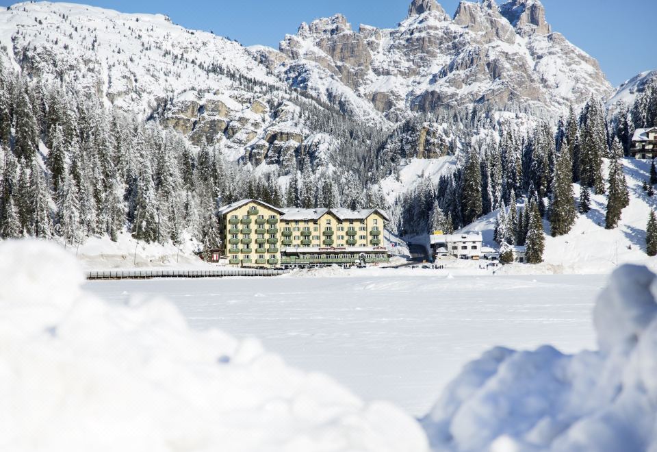 a snow - covered lake with a hotel in the background , surrounded by snow - covered mountains and trees at Grand Hotel Misurina