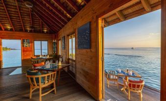 a wooden deck with a dining table and chairs , a window offering a view of the ocean , and lounge chairs at Komandoo Island Resort & Spa