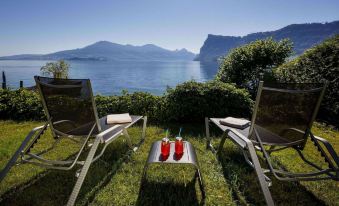a serene outdoor setting with two lounge chairs , a table , and two red objects on grass near the water 's edge at Seehotel Kastanienbaum