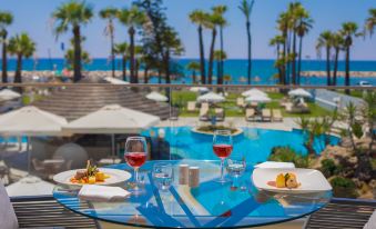 a dining table with two wine glasses and a plate of food , set on a balcony overlooking a swimming pool and palm trees at Golden Bay Beach Hotel