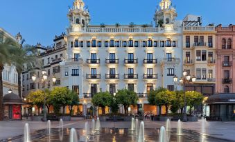 a city square with a fountain in the center , surrounded by tall buildings and a clock tower at H10 Palacio Colomera