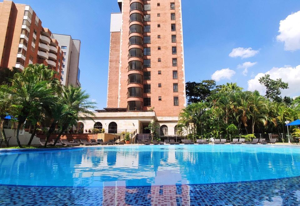 a tall building is reflected in a blue pool with palm trees in the background at Hotel Dann Carlton Medellín