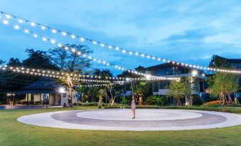 a woman is standing in a circle of lights on a grassy field , surrounded by buildings at Na Tree Tara Riverside Resort Amphawa Damnoensaduak