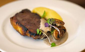 a white plate with a piece of meat and a side dish , including mushrooms , on a wooden table at The Cambridge Hotel