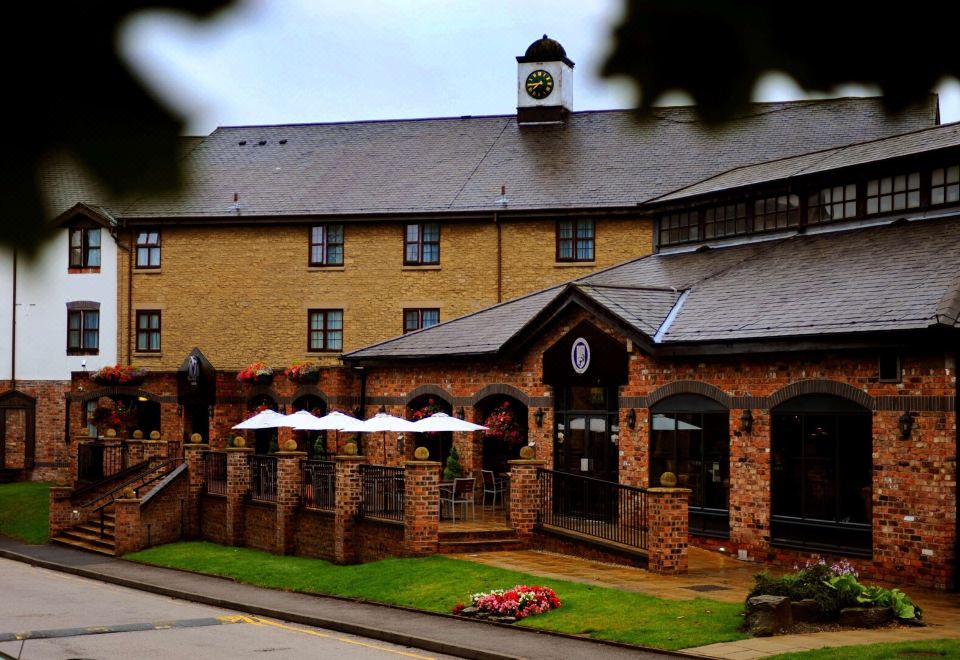 a brick building with a clock tower and umbrellas on the sidewalk , surrounded by greenery at Village Hotel Liverpool