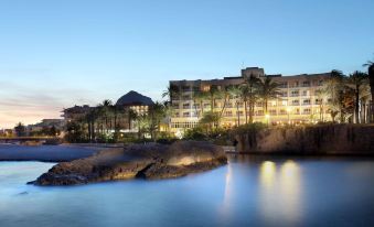 a large hotel surrounded by palm trees and water , with the sun setting in the background at Parador de Javea