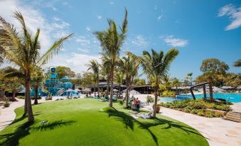 a lush green lawn with palm trees and people enjoying the water park in the background at Big4 Sandstone Point Holiday Resort Bribie Island