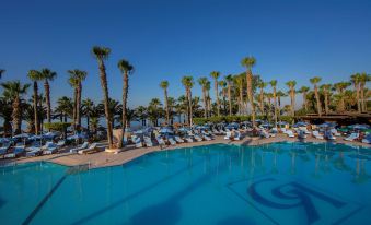 a large outdoor swimming pool surrounded by palm trees , with lounge chairs and umbrellas placed around the pool area at GrandResort by Leonardo Hotels