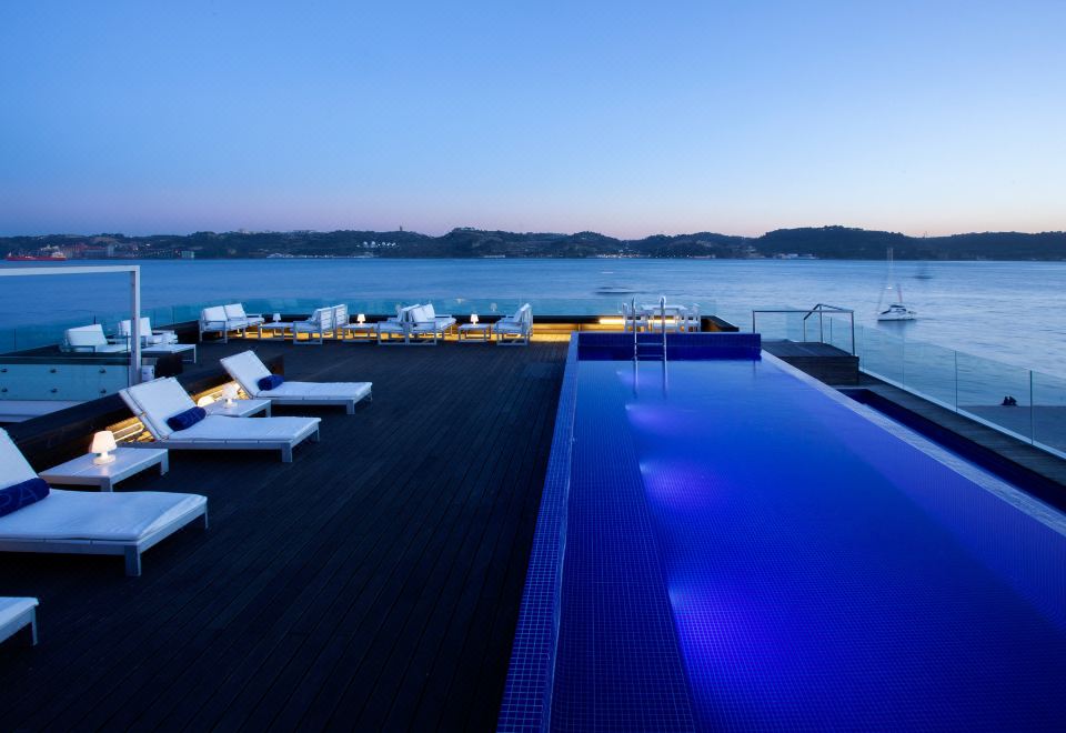 a large outdoor pool is surrounded by lounge chairs and a dock with boats in the background at Altis Belem Hotel & Spa, a Member of Design Hotels