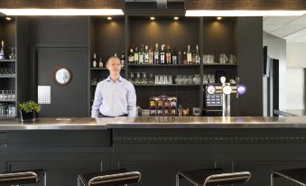 a man standing behind a bar , surrounded by various bottles and glasses , in a restaurant setting at Ibis Luxembourg Sud