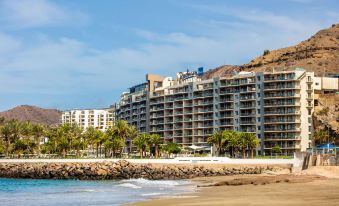 a beach with palm trees and a rocky shoreline , leading up to a multi - story building in the background at Radisson Blu Resort, Gran Canaria