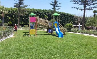 a colorful playground with a slide and swings is set in a grassy area surrounded by trees at Hotel Flora