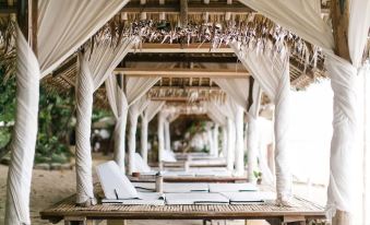 a row of white lounge chairs under a canopy , with white curtains hanging from the ceiling at La Luz Beach Resort