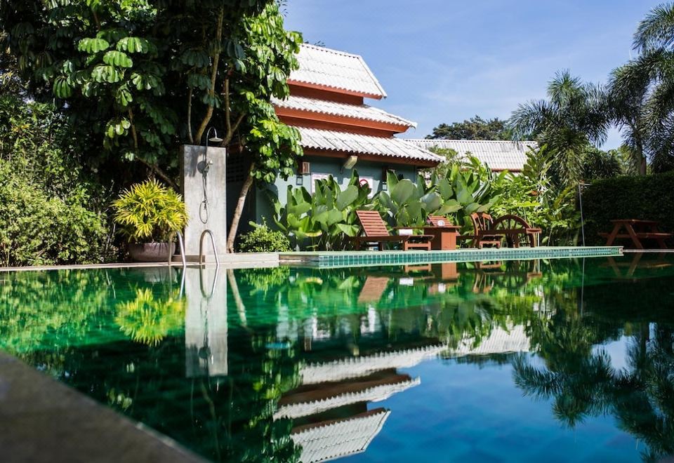 a large swimming pool surrounded by lush greenery , with a house in the background reflecting in the water at Poonyamantra Resort