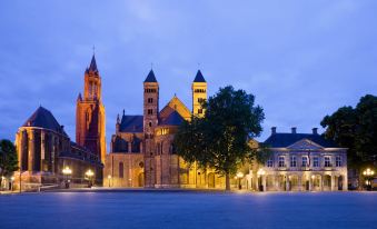 a large , ornate building with multiple spires is illuminated at night , surrounded by a plaza at Novotel Maastricht