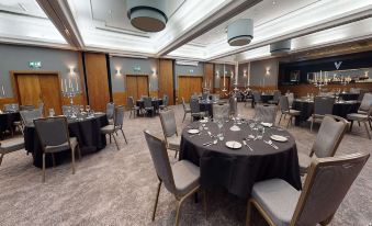 a large , empty banquet hall with multiple tables set for a formal event , surrounded by chairs and chandeliers at Village Hotel Chester St David's