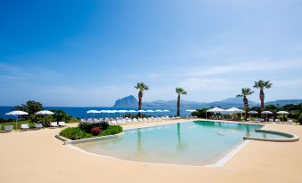 a large outdoor swimming pool surrounded by lounge chairs and umbrellas , with palm trees in the background at La Tonnara di Bonagia Resort