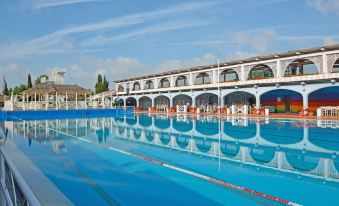 a large swimming pool with clear blue water , surrounded by white buildings and umbrellas , under a sunny sky at Hotel Boccaccio