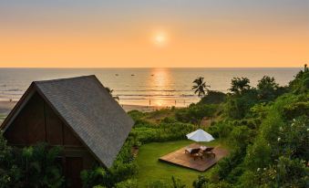 a beautiful beach house surrounded by lush green grass , with a sunset view of the ocean in the background at W Goa