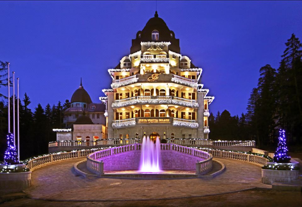 a grand building with multiple floors , lit up at night , and a large fountain in front of it at Festa Winter Palace Hotel