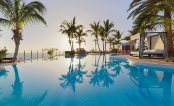 a large outdoor swimming pool surrounded by palm trees , with the sun setting in the background at Adrián Hoteles Roca Nivaria