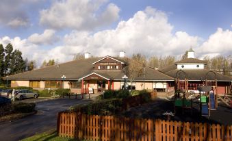 a large building with a red roof and several smaller buildings in the background , surrounded by trees at Premier Inn Liverpool (Tarbock)
