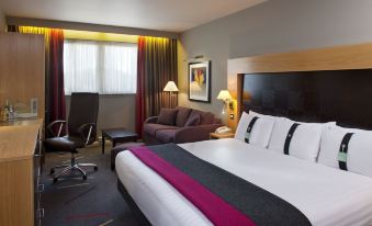 a large bed with a purple blanket is in the middle of a room with a chair , lamp , and window at Holiday Inn Aberdeen - West
