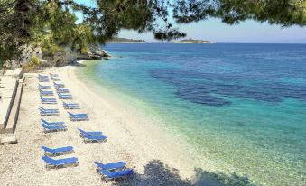 a beach with rows of blue chairs and umbrellas , set against the backdrop of a clear blue sea and trees at Agali Hotel Paxos