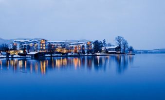 a large building with a clock on its facade is reflected in the water of a lake at Althoff Seehotel Überfahrt