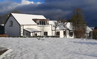 a snow - covered yard with a white house surrounded by trees and a cloudy sky in the background at Trenewydd Farm Holiday Cottages