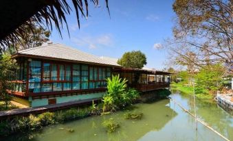 a serene view of a building surrounded by greenery and a pond , with clear blue skies above at Maikaew Damnoen Resort