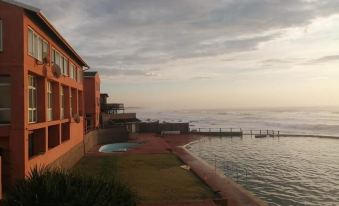 a beautiful sunset over the ocean , with a pool and building in the foreground , while the sun sets behind the water at Umkomaas Lodge