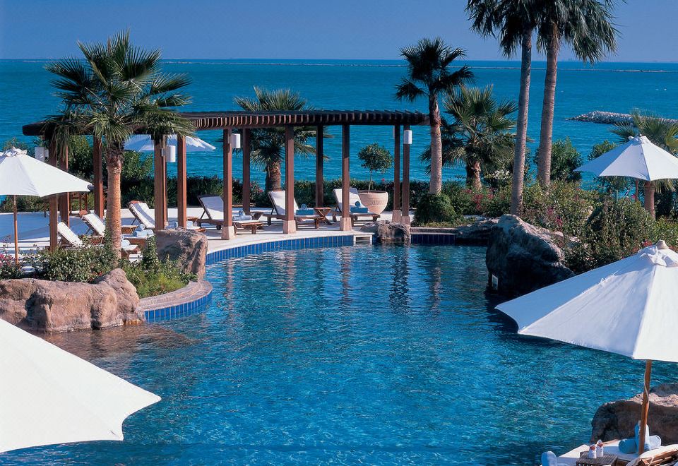 a large outdoor pool surrounded by lounge chairs and umbrellas , with palm trees in the background at The Ritz-Carlton, Doha