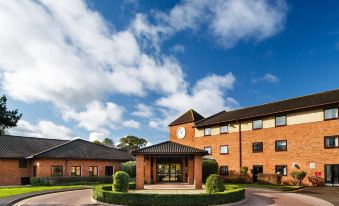 a brick building with a clock on the front , surrounded by trees and a blue sky at Delta Hotels Milton Keynes
