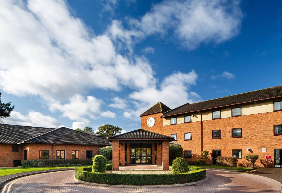 a brick building with a clock on the front , surrounded by trees and a blue sky at Delta Hotels Milton Keynes