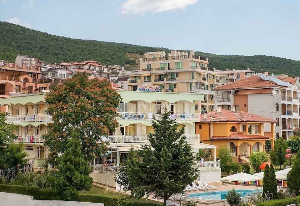 a sunny day in a resort with a large building , swimming pool , and trees , set against a backdrop of mountains at Hotel Panorama