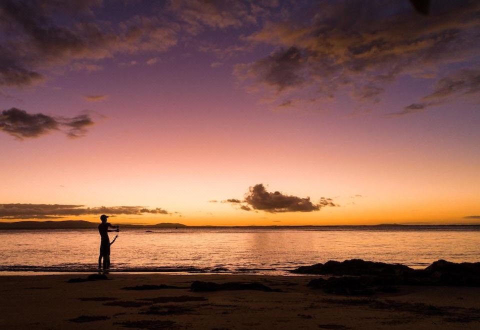 a person standing on the shore of a beach at sunset , casting a fishing rod at 1770 Beach Shacks