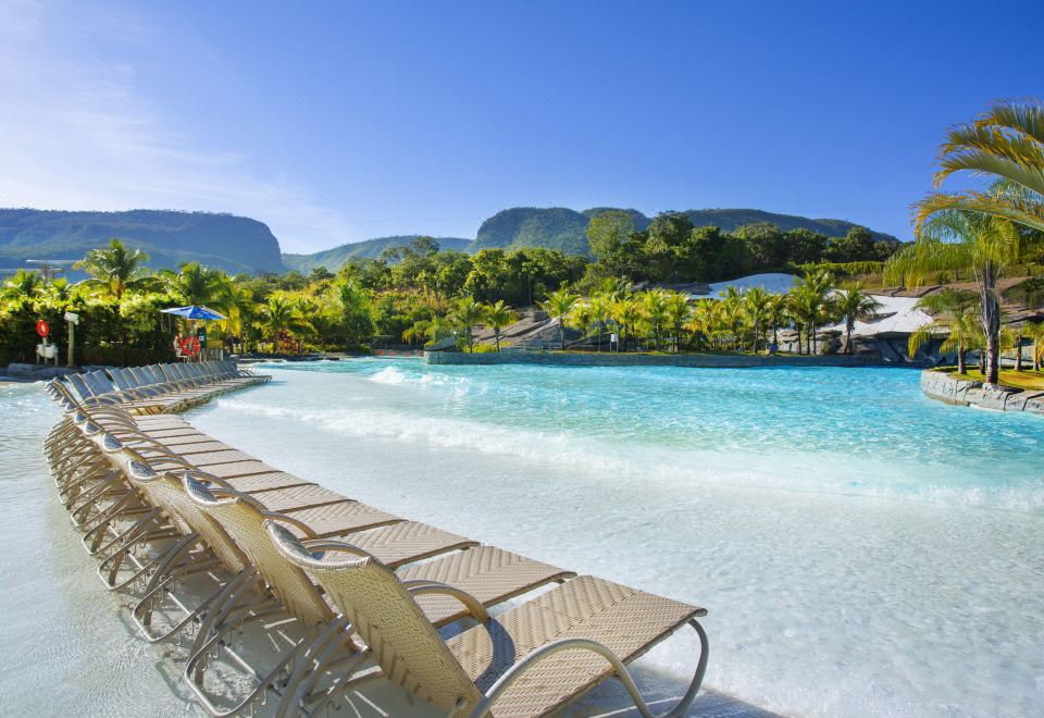 a large outdoor swimming pool surrounded by lounge chairs and umbrellas , with mountains in the background at Rio Quente Resorts - Hotel Pousada