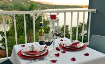 a dining table set with wine glasses , plates , and utensils on a balcony overlooking the water at The Shore House