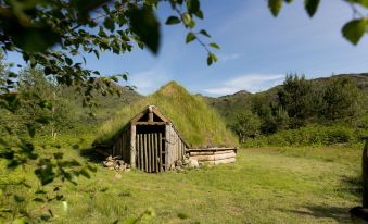 a wooden hut with a thatched roof is nestled in a green field with mountains in the background at YHA Eskdale