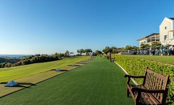 a long , grassy walkway with benches and chairs leading up to a golf course , surrounded by trees and buildings at Finca Cortesin Hotel Golf & Spa