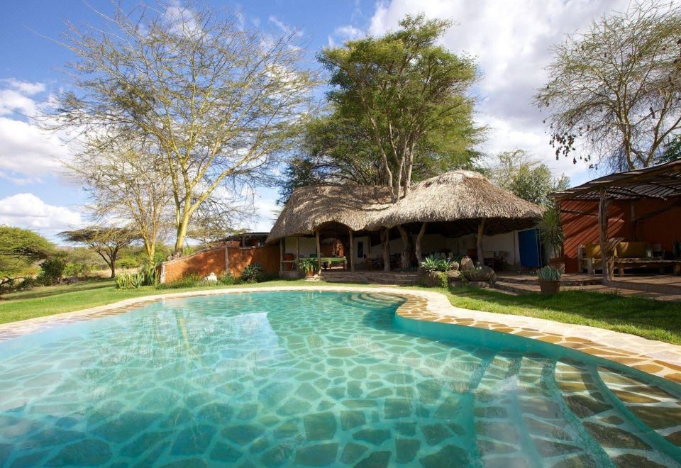 a large , clear swimming pool surrounded by lush green trees and a thatched - roof hut in the background at Elewana Lewa Safari Camp